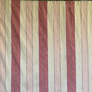 May include: Close-up of a fabric with vertical stripes. The stripes alternate between a muted red, off-white, and a thin black and brown stripe. The fabric has a slightly wrinkled texture, suggesting it is made of a natural fiber like cotton or linen.