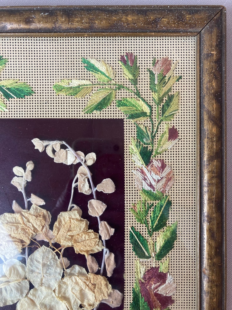 Op de afbeelding: Een ingelijst stuk geperste botanische kunst met gedroogde bladeren en bloemen. De lijst is van bruin hout met een gouden rand. Het kunstwerk bevindt zich achter een stuk gaas.
