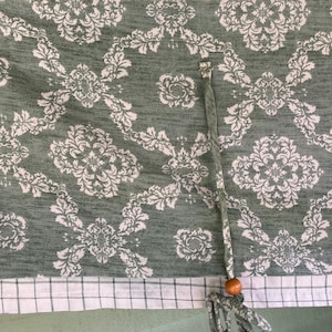 May include: A green and white patterned fabric with a damask design. The fabric features a repeating floral motif in white against a green background. A drawstring with a wooden bead is attached to the fabric.