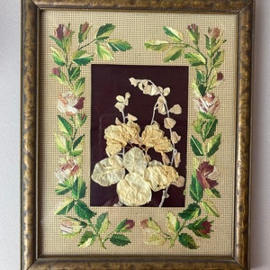 Op de afbeelding: Een ingelijst gedroogd bloemenkunstwerk met een bruine lijst en een beige achtergrond. De bloemen zijn geperst en gemonteerd op een donkerrode achtergrond. De lijst heeft een decoratieve rand van geborduurde bloemen.