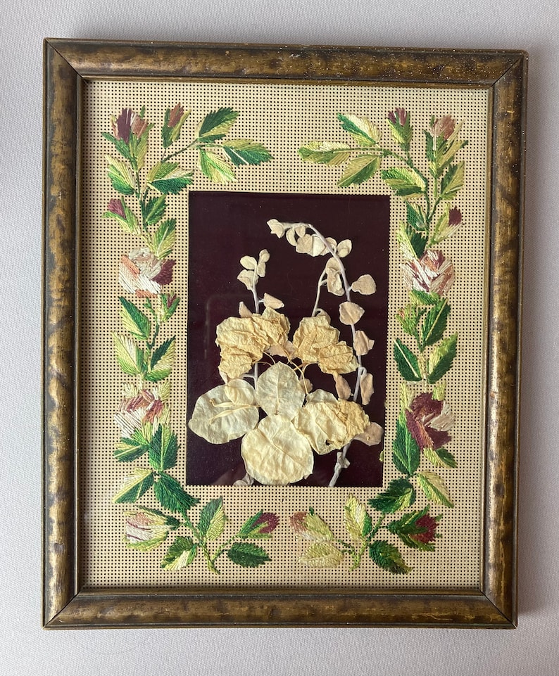Op de afbeelding: Een ingelijst gedroogd bloemenkunstwerk met een bruine lijst en een beige achtergrond. De bloemen zijn geperst en gemonteerd op een donkerrode achtergrond. De lijst heeft een decoratieve rand van geborduurde bloemen.