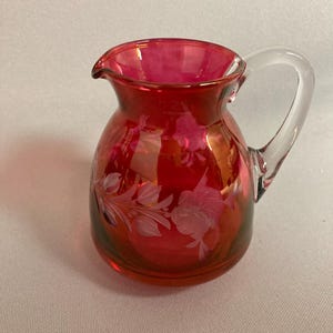 May include: A small, ruby red glass pitcher with a clear glass handle. The pitcher has a floral design etched into the glass.
