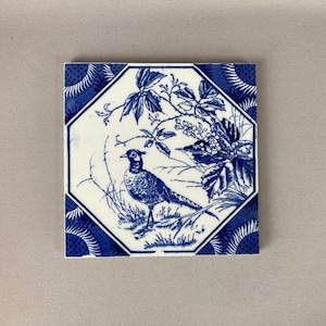 May include: Blue and white ceramic tile with a decorative design of a pheasant standing in a field of flowers and leaves.