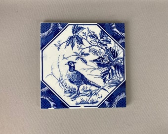 May include: Blue and white ceramic tile featuring a decorative design of a pheasant standing in a field of flowers and leaves.