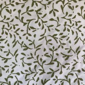 May include: White fabric with a repeating pattern of olive green leaves and vines. The leaves are stylized and arranged in a dense, organic design, creating a textured appearance. The fabric appears to be cotton or a similar material.