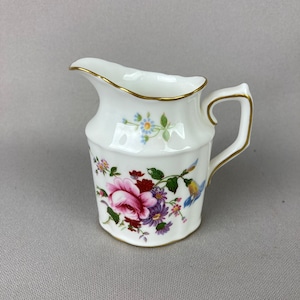 May include: A white porcelain creamer with a gold rim and floral pattern. The creamer features a pink rose, purple flowers, and blue birds.