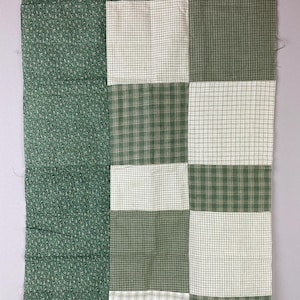 May include: A patchwork textile featuring a combination of green fabrics. The design includes a section of small, repeating patterns and a section of alternating squares of plaid and solid green. The textile is likely for home decor.