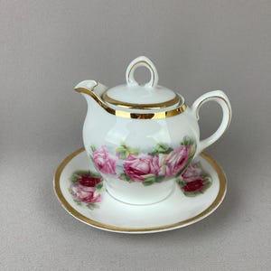 May include: A white porcelain tea set featuring a small teapot and saucer. The teapot and saucer are decorated with pink roses and gold trim. The teapot has a rounded shape with a handle and spout.