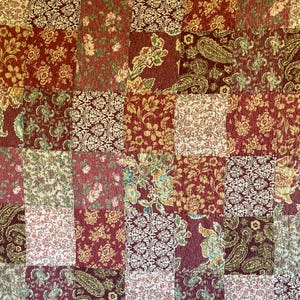 May include: A patchwork quilt featuring a grid of square fabric panels in shades of deep red, cream, and olive green. The fabric squares display various repeating patterns including floral motifs, intricate paisley swirls, and damask designs.