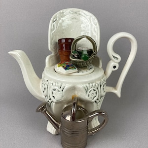 May include: White ceramic teapot shaped like a chair with a green and brown basket, a brown pot, and a green watering can. The teapot has a white handle and a spout.