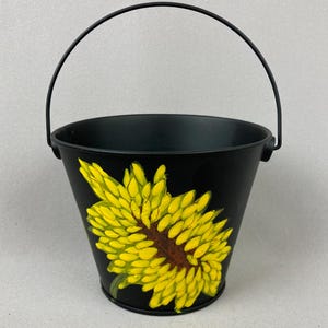May include: A black metal bucket with a thin, arched handle. The exterior features a painted design of a large, vibrant yellow flower with a dark brown center, showcasing detailed petals that extend across the front of the container.