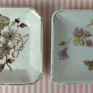 May include: Two small, square, white ceramic dishes with floral designs. One dish features a large flower with purple and gray accents, while the other has a butterfly and delicate pink and yellow flowers. Both have gold trim.