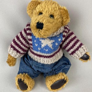 May include: A plush teddy bear with golden fur, wearing a blue and white star-patterned sweater and blue denim pants. The bear has a black nose and paws, and is sitting down.