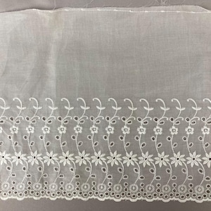 May include: White eyelet fabric with a floral design. The fabric features a delicate pattern of embroidered flowers and leaves along the bottom edge. The eyelet design includes small, circular cutouts, adding texture and detail to the textile.