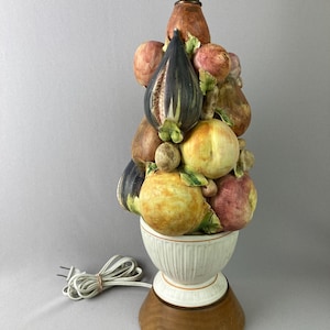 May include: A decorative table lamp featuring a stacked arrangement of ceramic fruits, including figs, peaches, and pears. The lamp has a white base with a wooden bottom and a brass-colored neck. The lamp is plugged into an electrical outlet.