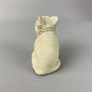 Pu&ograve; includere: Una piccola statuetta di ceramica bianca di un cane seduto con la testa bassa. Il cane indossa un collare con un design decorativo.