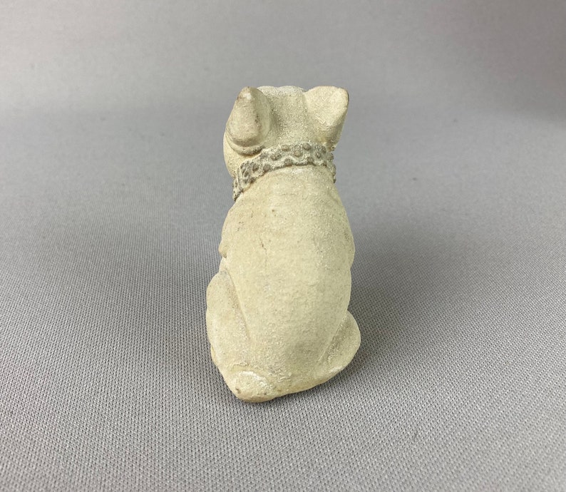 Pu&ograve; includere: Una piccola statuetta di ceramica bianca di un cane seduto con la testa bassa. Il cane indossa un collare con un design decorativo.