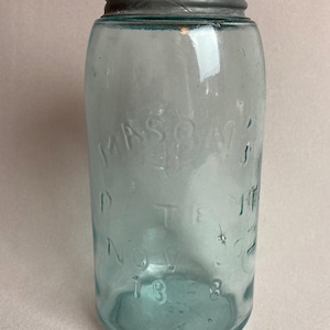 May include: A vintage blue-tinted glass Mason jar with a metal lid. The jar has embossed text that reads "MASON" and "1883". The jar is cylindrical with a slightly rounded shoulder and a threaded neck for the lid.