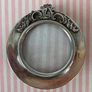 May include: A silver-toned round picture frame with an ornate design around the edge. The frame has a small, clear glass insert for a photo.
