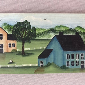 May include: A hand-painted wooden sign depicting a rural scene with two houses. The house on the left is yellow with a black door and windows. The house on the right is blue with a black roof and white windows. Both houses are surrounded by green grass and a white picket fence.