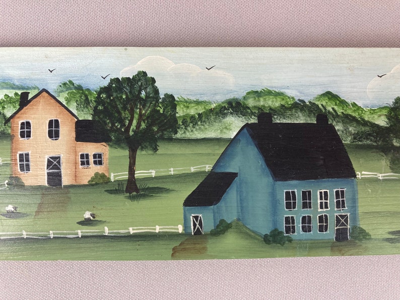 May include: A hand-painted wooden sign depicting a rural scene with two houses. The house on the left is yellow with a black door and windows. The house on the right is blue with a black roof and white windows. Both houses are surrounded by green grass and a white picket fence.
