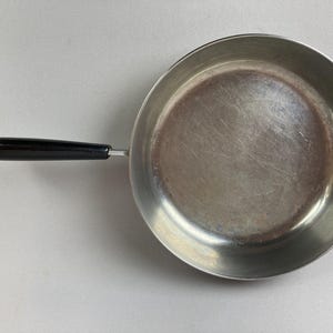 May include: A stainless steel frying pan with a black handle. The pan has a circular cooking surface and a long, curved handle. The handle is made of a dark, glossy material. The pan is designed for cooking.