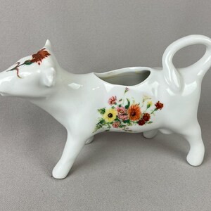 May include: White ceramic cow creamer with a floral design. The creamer has a handle shaped like a tail and a spout on the back. The cow has a floral design on its side and a brown and green floral design on its head.