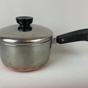 May include: A small, stainless steel saucepan with a lid and a black handle. The pot has a copper-colored bottom and a black knob on the lid. The handle has a metal loop at the end.