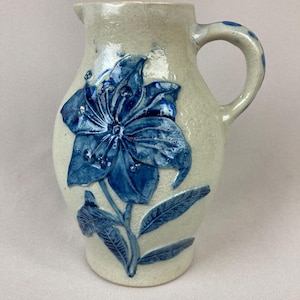 May include: A stoneware pitcher with a light beige background and a large blue floral design. The pitcher has a handle and a spout. The floral design is a raised relief, adding texture to the piece. The pitcher is likely handmade.
