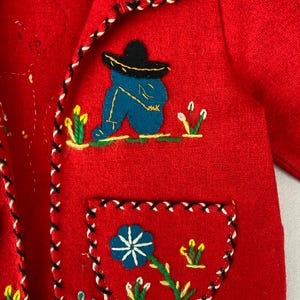 以下が含まれることがあります： 赤いウールジャケットにカラフルな刺繍が施されています。ジャケットには、ソンブレロをかぶった男性、花、緑の葉が描かれています。ジャケットの縁には黒と白のトリムが施されています。