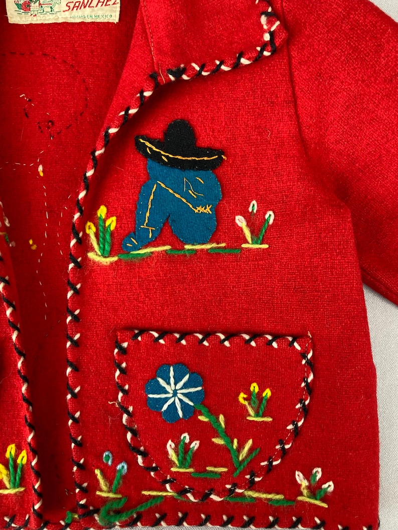 以下が含まれることがあります： 赤いウールジャケットにカラフルな刺繍が施されています。ジャケットには、ソンブレロをかぶった男性、花、緑の葉が描かれています。ジャケットの縁には黒と白のトリムが施されています。