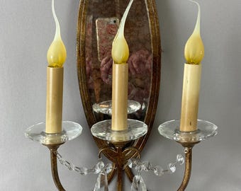 May include: An antique wall sconce with an oval mirror and three candle holders. The sconce has a gold finish with clear crystal accents and faux candle lights with yellow flames. The mirror has a distressed finish.