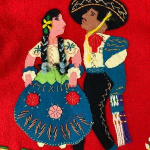 以下が含まれることがあります： 伝統的なメキシコの衣装を着た男性と女性の刺繍が施されたフェルトの壁掛け。女性は赤い花がついた青いスカートを、男性は青いジャケットと黒いズボンを着用しています。背景は赤で、女性のスカートには「MEXICO」という文字が刺繍されています。