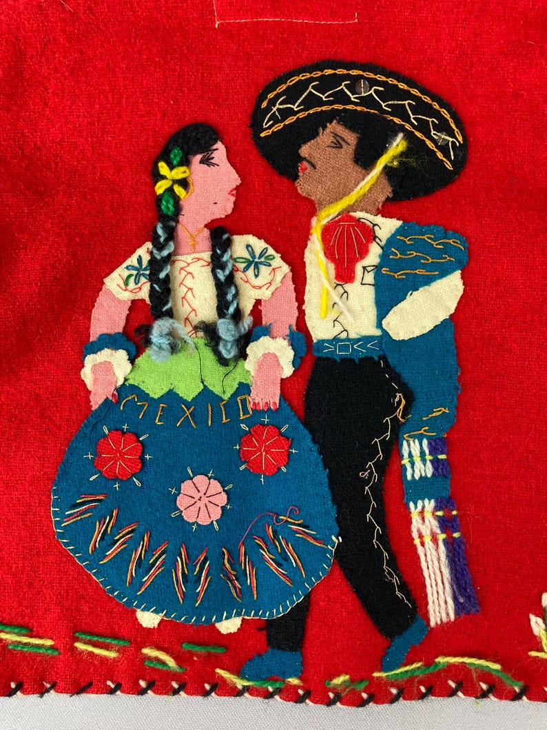 以下が含まれることがあります： 伝統的なメキシコの衣装を着た男性と女性の刺繍が施されたフェルトの壁掛け。女性は赤い花がついた青いスカートを、男性は青いジャケットと黒いズボンを着用しています。背景は赤で、女性のスカートには「MEXICO」という文字が刺繍されています。