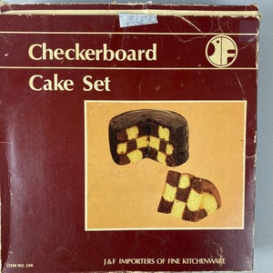 May include: A vintage Checkerboard Cake Set box with a picture of a chocolate and yellow checkerboard cake. The box is a deep red-brown color with the words "Checkerboard Cake Set" in a cream color.