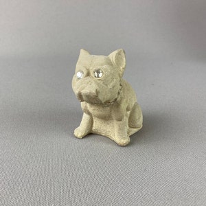 Pu&ograve; includere: Una piccola statuetta di ceramica grigia di un cane seduto con la testa inclinata di lato. Il cane ha due occhi di strass trasparenti.