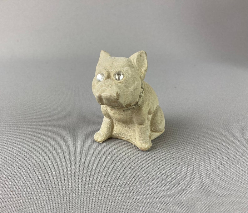 Pu&ograve; includere: Una piccola statuetta di ceramica grigia di un cane seduto con la testa inclinata di lato. Il cane ha due occhi di strass trasparenti.