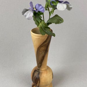 Op de afbeelding: Een hoge, slanke houten vaas met een ronde voet en een uitlopende opening. Het hout heeft een natuurlijke lichtbruine kleur met opvallende donkerbruine nerven. In de vaas staan twee kunstmatige viooltjes met paarse en witte bloemblaadjes.