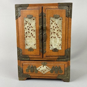 May include: A small, wooden cabinet with two doors and a drawer. The cabinet is a warm brown color with brass corner brackets and hinges. The doors feature carved, off-white panels. The drawer has decorative brass accents.
