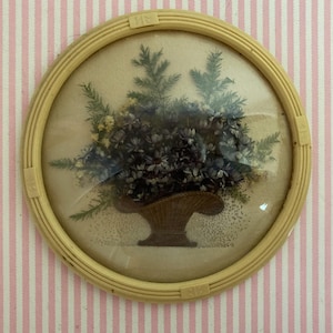 May include: A vintage floral arrangement in a round, cream-colored frame. The arrangement features blue and yellow flowers and green foliage in a woven basket. The frame is set against a pink and white striped background.