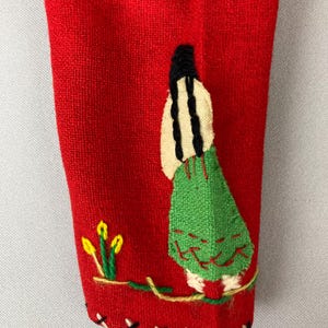 以下が含まれることがあります： 赤い布地に、長い黒髪の女性が緑色の刺繍で描かれている。女性は黄色の花が咲いた枝の上に立っている。