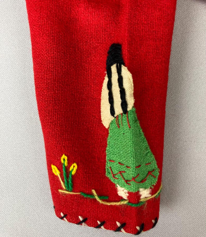 以下が含まれることがあります： 赤い布地に、長い黒髪の女性が緑色の刺繍で描かれている。女性は黄色の花が咲いた枝の上に立っている。