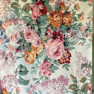 May include: Floral fabric with a cream background, featuring a pattern of large roses in shades of pink, orange, and red, interspersed with smaller lilac-colored flowers and green leaves. The fabric has a ruffled edge.