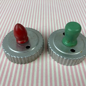 May include: Two vintage metal cookie cutters with wooden handles. The cutter on the left has a red handle and the cutter on the right has a green handle.
