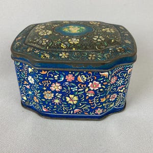 May include: A vintage blue metal box with a floral design. The box has a dark blue lid with a floral pattern and a lighter blue body with colorful flowers. The box is likely used for storing small items.