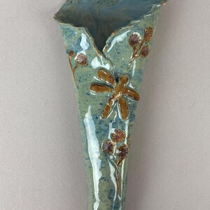 May include: A handmade ceramic wall vase in a muted blue-green glaze. The vase features a dragonfly and floral embellishments in brown. The vase is designed with a flared top and a narrow, elongated body, perfect for displaying flowers.