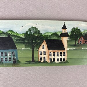 May include: A hand-painted wooden sign depicting a charming rural scene with three houses. The houses are painted in shades of blue, yellow, and red, with black roofs and white windows. The sign is set against a backdrop of green hills and trees.