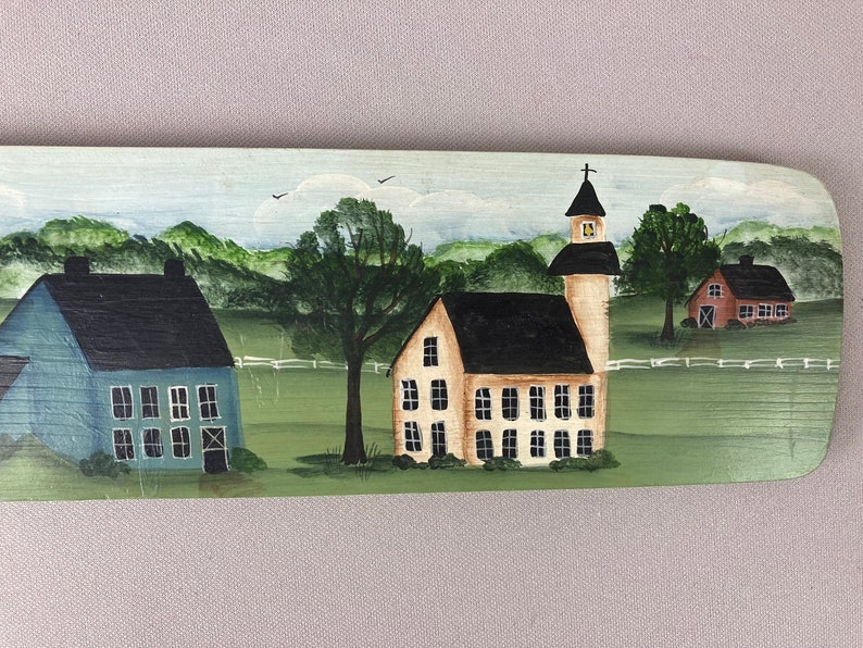 May include: A hand-painted wooden sign depicting a charming rural scene with three houses. The houses are painted in shades of blue, yellow, and red, with black roofs and white windows. The sign is set against a backdrop of green hills and trees.