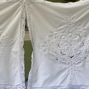 May include: Two white, embroidered curtains with floral and geometric cut-out designs. The curtains have a scalloped edge and are made of a soft, flowing fabric. The design features large, stylized flowers.
