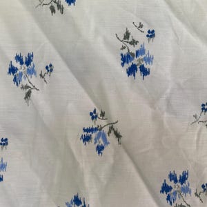 May include: White fabric with a repeating pattern of blue and gray floral designs. The flowers have a textured, ikat-like appearance. The fabric has subtle horizontal lines, adding to its visual texture. The overall aesthetic is soft and delicate.